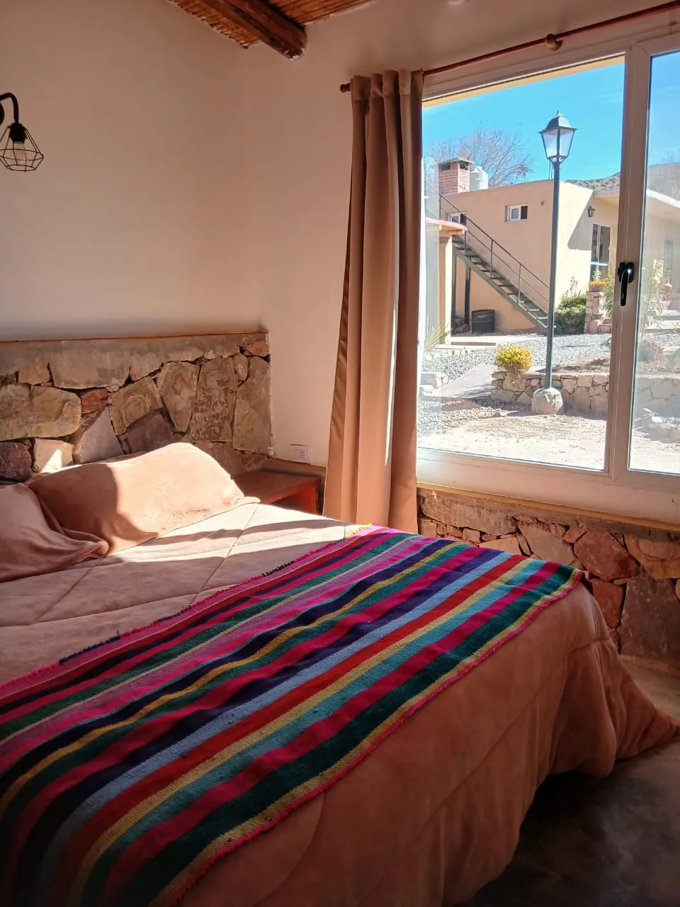 Cabaña Fuego: cama con manta de rayas de colores, pared de piedra y ventana al patio